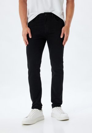 Jeans ajustados negros de mezclilla, con cierre de botón frontal, trabillas para cinturón y cinco bolsillos. Combinados con zapatillas blancas.