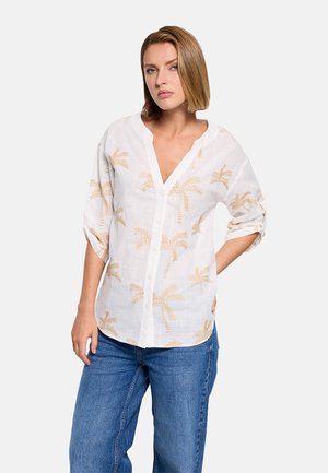 Mujer con blusa blanca abotonada con estampado de hojas de palma beige y jeans azules, de pie con una mano en la cadera contra un fondo blanco.