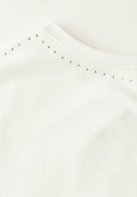 T-shirt en coton blanc avec un col rond. Des clous en argent décoratifs bordent la couture de l'épaule, ajoutant une touche de texture.