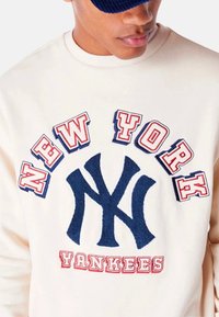 Sudadera de crema con el logo "NY" en azul marino y el texto "NEW YORK YANKEES" en rojo. Presenta bordado texturizado y un cuello redondo.