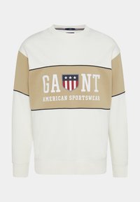 Sweatshirt crema con una striscia orizzontale beige. La scritta "GAUNT" e "AMERICAN SPORTSWEAR" è stampata in bianco. Presenta un logo a scudo con accenti rossi, bianchi e blu.