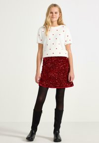 Hvid, fluffy sweater med røde hjerte-mønstre og korte ærmer, parret med en rød, pailletbesat nederdel. Sorte strømpebukser og knæhøje sorte støvler.