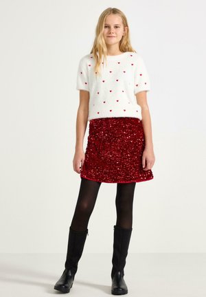 Hvid, fluffy sweater med røde hjerte-mønstre og korte ærmer, parret med en rød, pailletbesat nederdel. Sorte strømpebukser og knæhøje sorte støvler.
