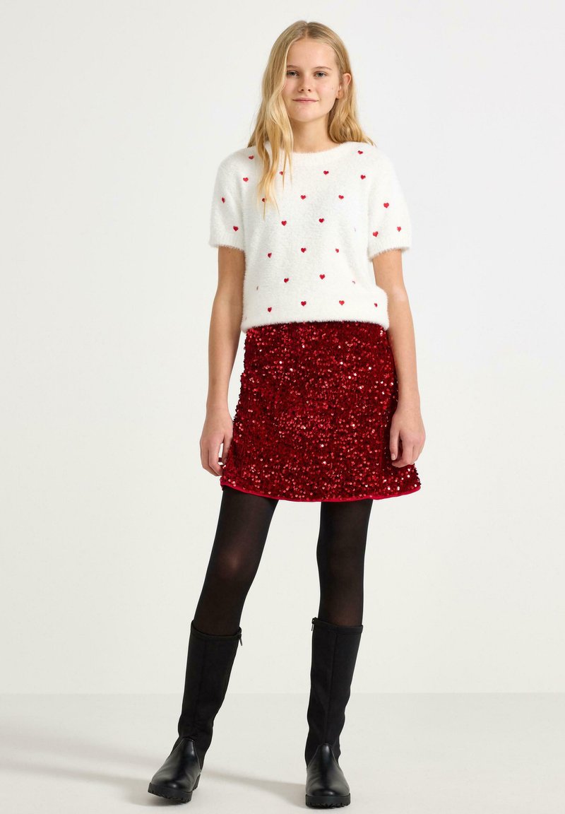 Hvid, fluffy sweater med røde hjerte-mønstre og korte ærmer, parret med en rød, pailletbesat nederdel. Sorte strømpebukser og knæhøje sorte støvler.