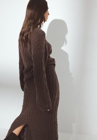 Vestido marrón de punto con mangas largas y un diseño texturizado y estampado. Presenta una apertura lateral para mayor movimiento y forma.