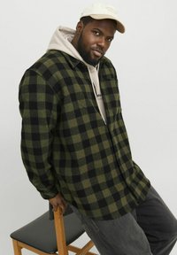 Zwart en groen geruite flanellen shirt met een zak; gedragen over een beige hoodie. Gekocht met grijze broek en een lichtgekleurde pet.