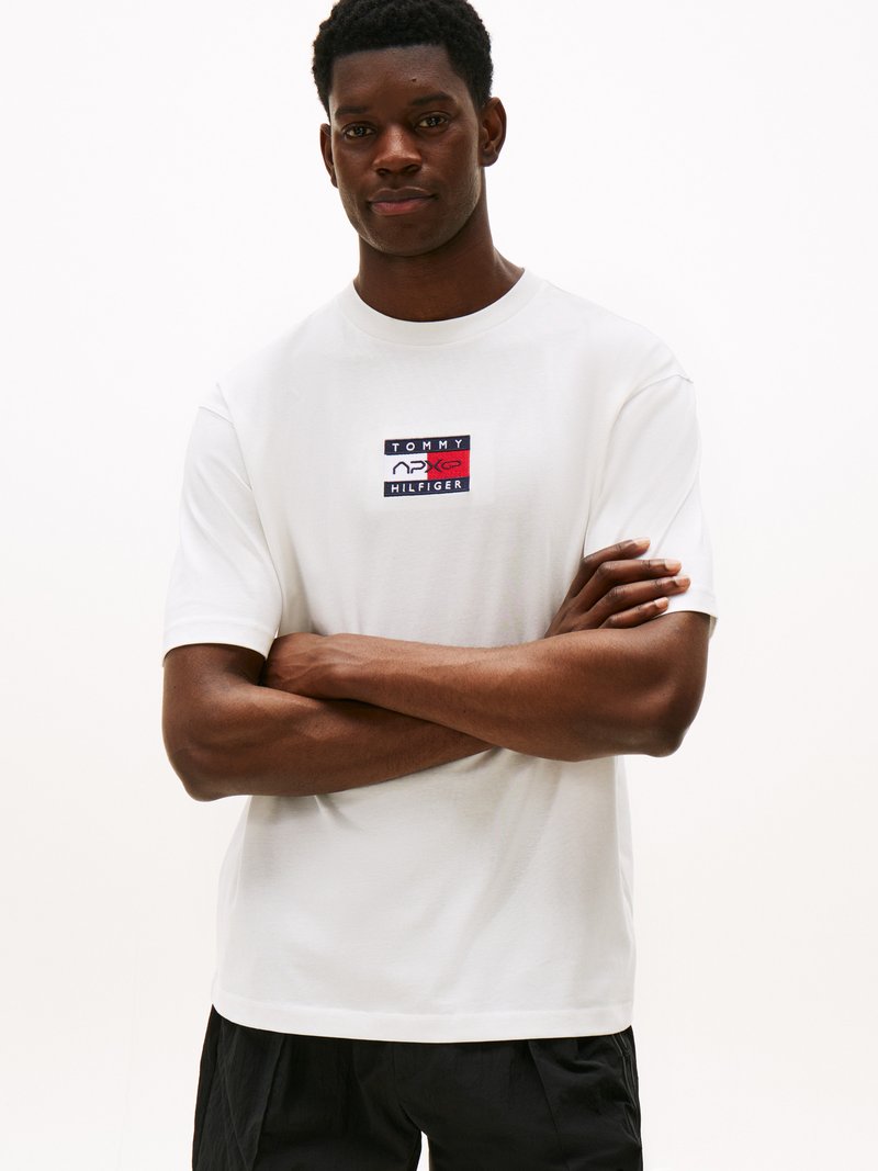 Baltas trumpas rankovėmis marškinėliai su stačiakampiu tamsiai mėlynos, raudonos ir baltos spalvos logotipo pleistru, ant kurio užrašyta „TOMMY HILFIGER“; švelni medvilnės tekstūra.