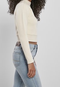 Top de manga larga color crema con cuello alto, tela texturizada y corte ajustado, combinado con jeans azul claro y diseño sutil en el bolsillo trasero.