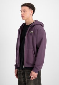 Fialová mikina na zips vyrobená z mäkkej látky, s prednými vreckami a kapucňou so šnúrkou. Pod ňou čierne tričko; model stojí v profilu.
