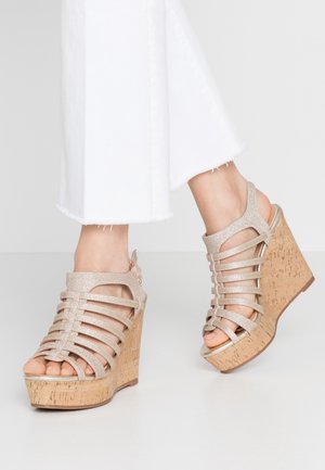 Füße, die beige, glitzernde Riemchen-Wedges mit Korkabsätzen tragen, kombiniert mit weißen, gekürzten Hosen.