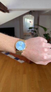 Gold-tone watch with a mesh band, blue dial, marked with silver hour and minute hands, square date window, and brand name "Guess."