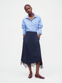 Femme portant une chemise boutonnée rayée bleue rentrée dans une jupe marine à ourlet en dentelle noire, associée à des chaussures Mary Jane rouges, debout les mains dans les poches.
