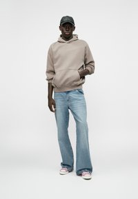 Μπεζ φόρμες με καγκουρό τσέπη, ανοιχτό μπλε φαρδιά τζιν και sneakers με ροζ λεπτομέρειες. Άνδρας στέκεται με το ένα χέρι στην τσέπη.