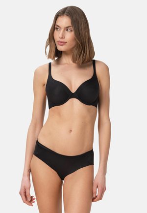 Femme aux cheveux bruns mi-longs portant un soutien-gorge noir et une culotte assortie, debout devant un fond clair uni.