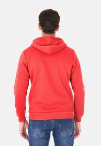 Felpa rossa in cotone con tasca a marsupio, polsini a costine e cappuccio con cordino. Indossata con jeans in denim blu, vista da dietro.