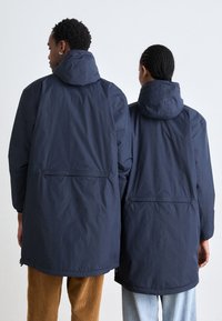 Vestes imperméables bleu marine avec capuches, dotées d'une poche arrière zippée, d'une coupe décontractée et d'une texture lisse, portées par deux personnes vues de dos.
