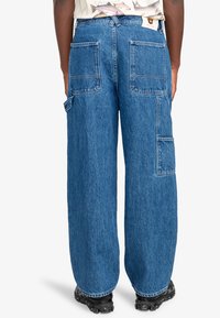 Jeans in denim blu a vestibilità ampia con diverse tasche applicate, cuciture visibili e una silhouette a gamba dritta. Presenta un vita alta e un tessuto robusto.