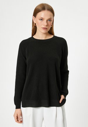 Schwarzer Strickpullover mit rundem Halsausschnitt und langen Ärmeln. Verfügt über eine gerippte Textur und eine lockere Passform, getragen über weißen Hosen.