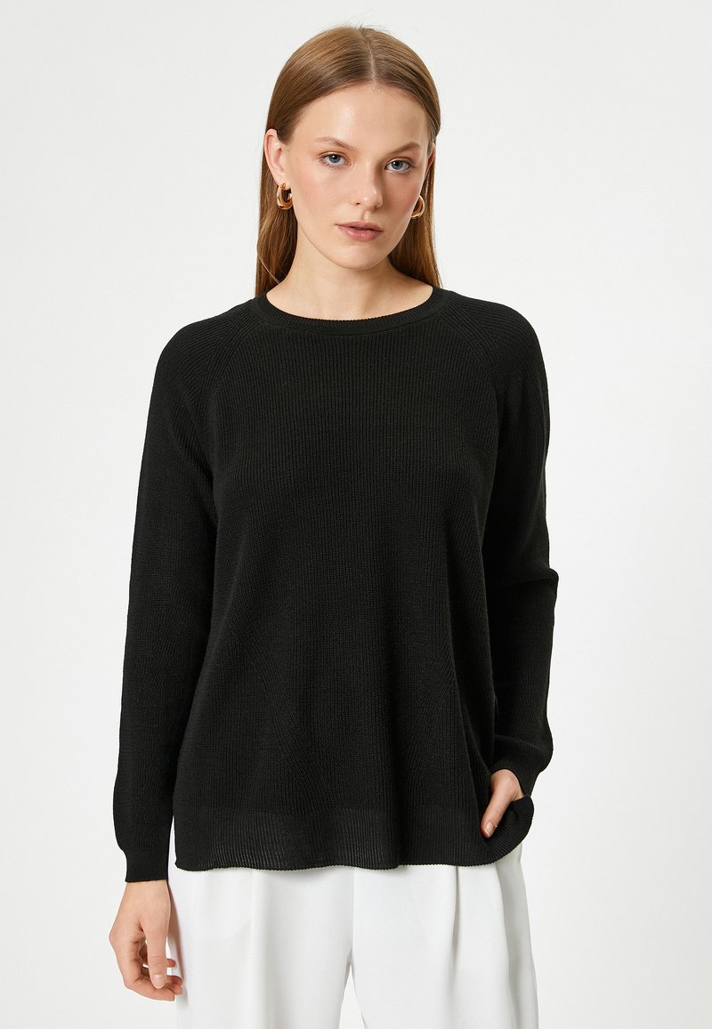 Maglione nero a maglia con scollo rotondo e maniche lunghe. Presenta una texture a coste e una vestibilità morbida, indossato sopra pantaloni bianchi.