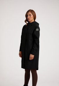 Zwarte waterdichte jas met een capuchon, voorzien van een voorrits, zijzakken en een logopatch op de linker mouw. Glad textuur, knielang.