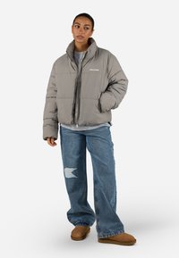 Grå pufferjakke med høy krage, glidelås foran og logo. Matchet med blå vide jeans med hull, og brune slip-on sko.