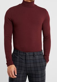 Bordowa bluzka z długim rękawem i golfem, dopasowana, z prążkowanym kołnierzem, zestawiona z ciemnymi spodniami w kratę, posiadającymi zapięcie na guziki i boczne kieszenie.