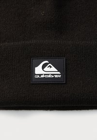 Fekete kötött sapka, amelyen elöl egy téglalap alakú fekete gumipárna található fehér logóval és "QUIKSILVER" felirattal.