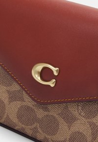 Červená kožená peněženka s klopou a hnědým vzorovaným plátnem. Má zlatý akcent s logem a žluté švy podél okraje pro zvýraznění detailu.