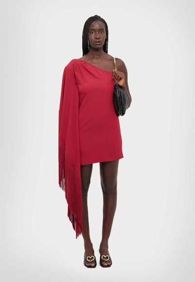 Vestido asimétrico rojo con un detalle de flecos en un lado, que cuenta con una sola tira. Bolso negro y sandalias deslizantes con acentos en forma de corazón.