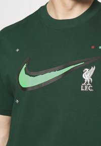 Ciemnozielona koszulka bawełniana z dużym czarnym i neonowozielonym logo Nike w formie "swoosh" oraz z logo Liverpool F.C. na lewej piersi.