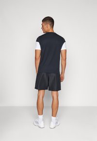 Dunkelblaues kurzärmliges Hemd mit weißen Akzenten, kombiniert mit schwarzen Shorts und weißen Sportschuhen, von hinten gezeigt. Glatte Stoffoberfläche.