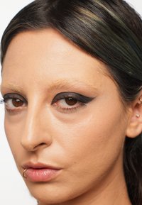 Close-up van een gezicht met een gladde, lichtbruine huid, dramatische zwarte eyeliner en een gouden lippiercing. Donker haar met groene accenten.