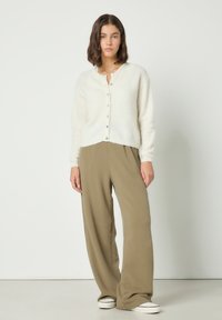 Mlada ženska, oblečena v belo džemper s gumbi, široke rjave hlače in bele superge, stoji pred enobarvnim belim ozadjem.