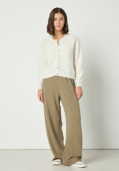Jeune femme portant un cardigan blanc boutonné, un pantalon marron ample et des baskets blanches, debout devant un fond blanc uni.