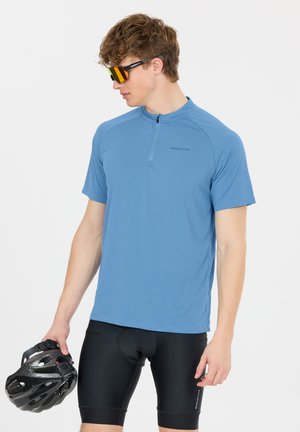 Junger Mann in blauem Radtrikot, schwarzen gepolsterten Shorts und reflektierenden Sonnenbrillen, hält einen schwarzen Fahrradhelm und blickt leicht nach links.
