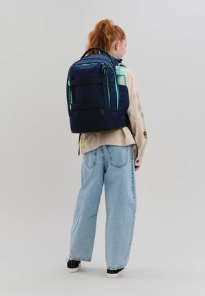Junge Person mit roten Haaren, die weite Jeans, eine beige Jacke und einen großen dunkelblauen Rucksack trägt und eine mintgrüne Wasserflasche hält.