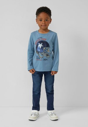 Lichtblauw langemouw shirt met een tijgerafbeelding en pailletten, gecombineerd met donkerblauwe spijkerbroek en witte sneakers, tegen een neutrale achtergrond.