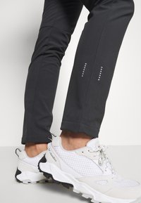 Persona vestida con pantalones deportivos negros con puntos reflectantes y zapatillas de senderismo blancas de Columbia, de pie sobre un suelo blanco contra una pared blanca.