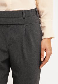 Pantalons gris ajustés avec une texture lisse, présentant un devant plat, des passants de ceinture et des poches latérales, associés à un haut beige clair.