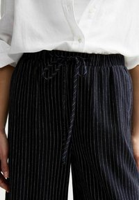 Pantalon rayé marine en tissu doux avec une taille élastique et un cordon tressé. Rayures blanches verticales et poches latérales visibles.