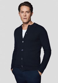 Męski ciemnoniebieski kardigan, dzianinowa faktura, zapinany na guziki, z dwoma guzikami i płaskim kołnierzem. Nosi go na białej koszuli.