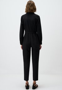 Zwarte jumpsuit met lange mouwen, fitted taille en rechte pijpen. Soepele stof met een subtiele glans. Verstelbare riem in de taille.