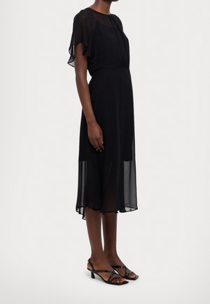 Femme portant une robe midi noire transparente avec des manches flottantes et des sandales noires à talons bas et à brides, debout devant un fond uni.