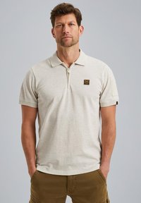 Polo shirt beige clair à manches courtes avec un col zippé, un patch sur la poitrine gauche et un tissu texturé. Les poignets ont de petits accents.