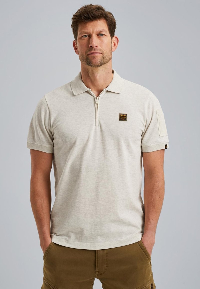 Polo shirt beige clair à manches courtes avec un col zippé, un patch sur la poitrine gauche et un tissu texturé. Les poignets ont de petits accents.