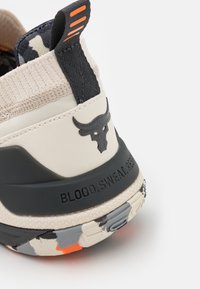 Beżowy but sportowy z czarnymi akcentami, teksturowaną tkaniną przy kołnierzu, podeszwą w kamuflaż oraz wypukłym emblematem byka. Napis brzmi "KREW, POT, SZACUNEK."