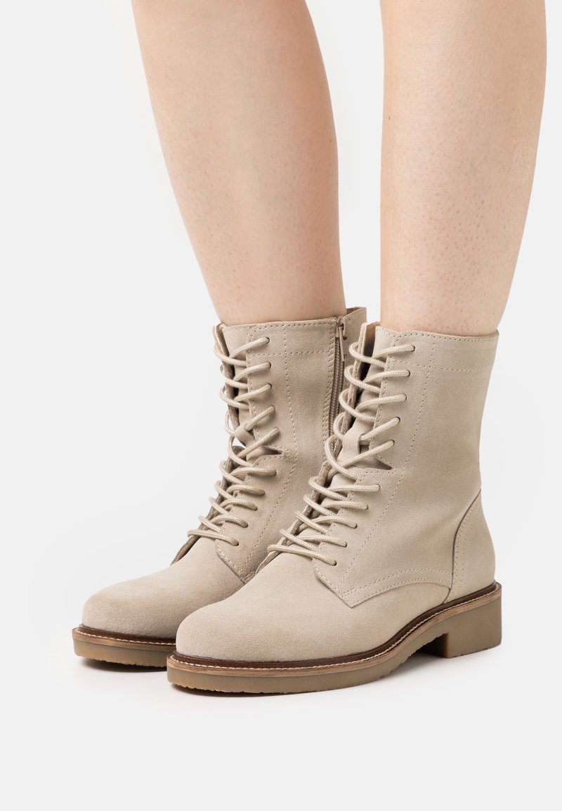 Bottines en daim beige avec lacets à l'avant, fermeture éclair sur le côté, petit talon et bout rond, portées sur des jambes nues sur un fond blanc.