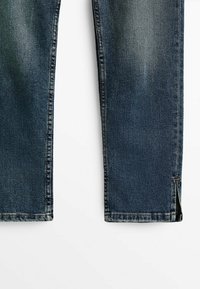Jean en denim bleu foncé avec une texture délavée, jambes droites, coutures contrastées et fentes sur les côtés à l'ourlet pour plus de détails.