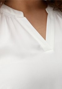 Witte blouse met een V-hals en rimpeldetails op de schouders, gemaakt van gladde stof met een zachte textuur.
