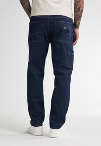 Jeans in denim blu scuro con vestibilità rilassata, dotati di due tasche posteriori e dettagli di cucitura visibili, abbinati a sneaker bianche.